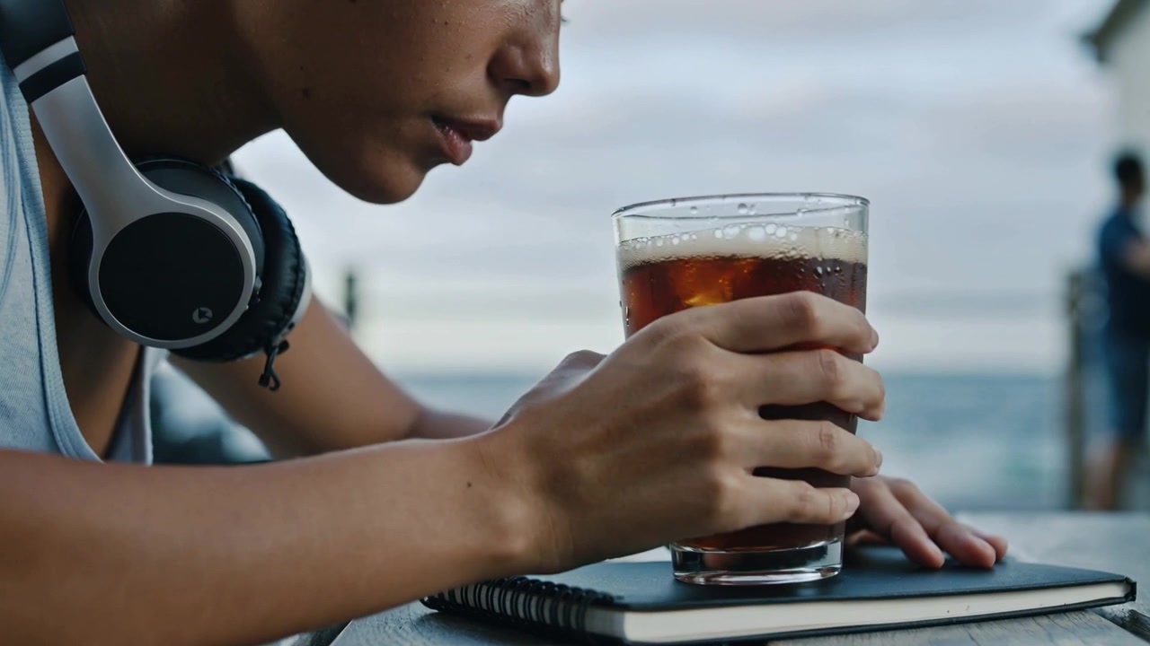 Kvinne i ermeløs topp med hodetelefoner lener seg og strekker seg etter et glass ved et bord ved bryggen, med en notatbok.