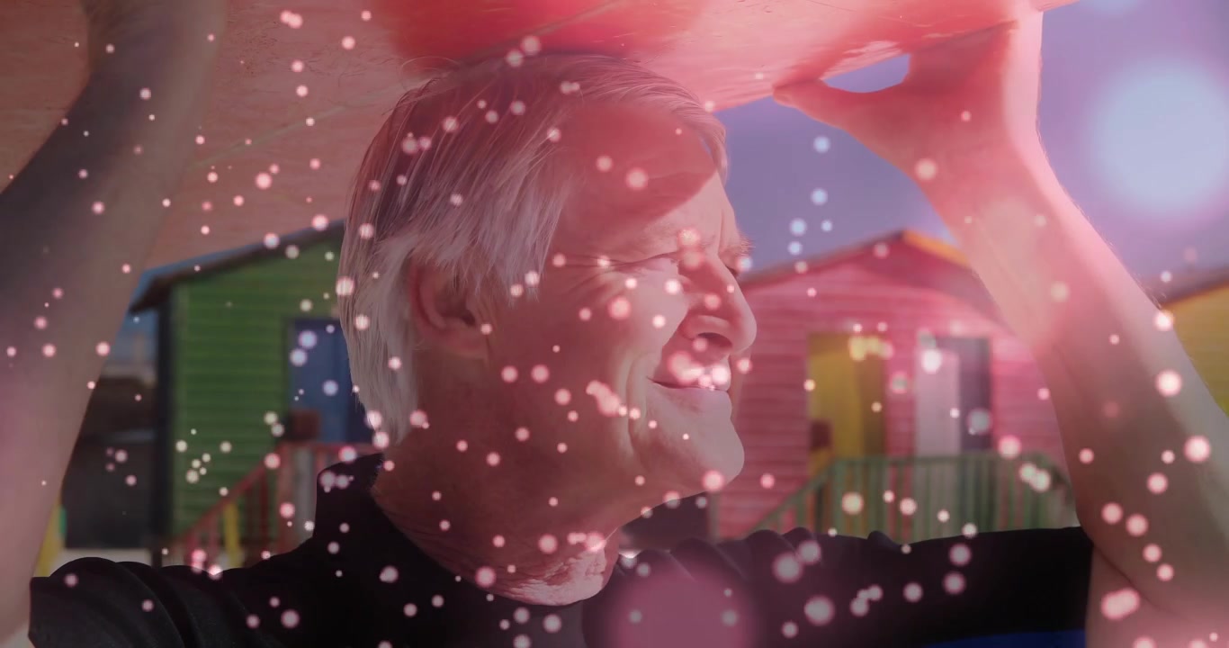 Holding pink shade, senior in dark top squinting with sunlight hitting face, bokeh at beach huts
