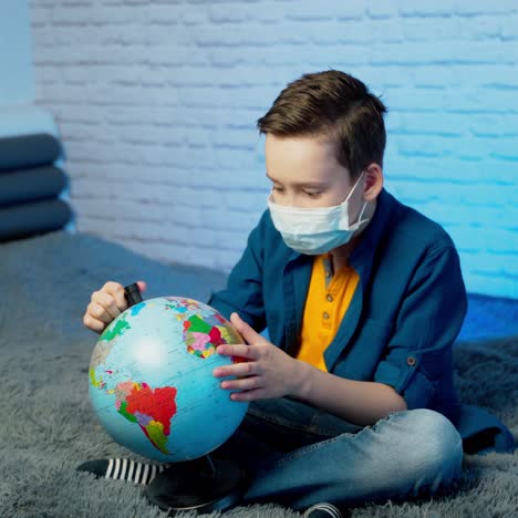 child in self isolation looking at globe learning about covid-19. boy in a medical mask with curious face thinking about the planet during covid lock down, quarantine and social distancing concept
