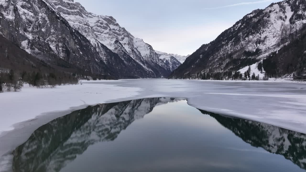 View of the icy surface of Klöntalersee in Klöntal, Switzerland, with snow-covered mountains towering over the frozen lake. The tranquil reflection of the landscape creates a serene winter scene.