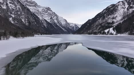 View of the icy surface of Klöntalersee in Klöntal, Switzerland, with snow-covered mountains towering over the frozen lake. The tranquil reflection of the landscape creates a serene winter scene.