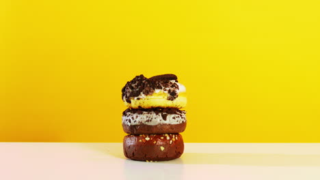 a hand uses a fork to pick up the top donut from a visually appealing stack of three donuts, all set against a vibrant yellow background, highlighting textures and toppings.