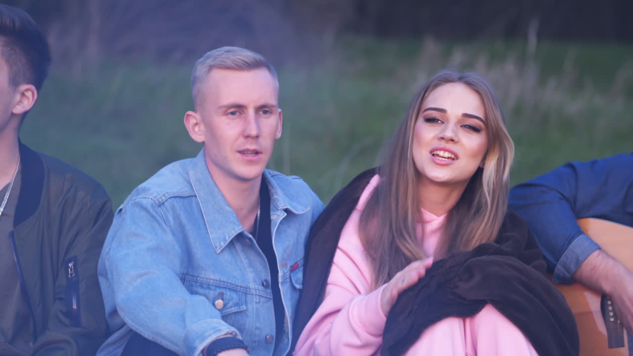 Young couple sitting in the centre and singing. Dating pair in the company of friends having picnic together outside city.