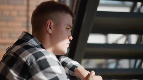 side profile of man sitting on metal staircase railing smoking cigarette in deep thought exhaling wisps of smoke into warm sunlight casting brick wall shadows in quiet urban setting