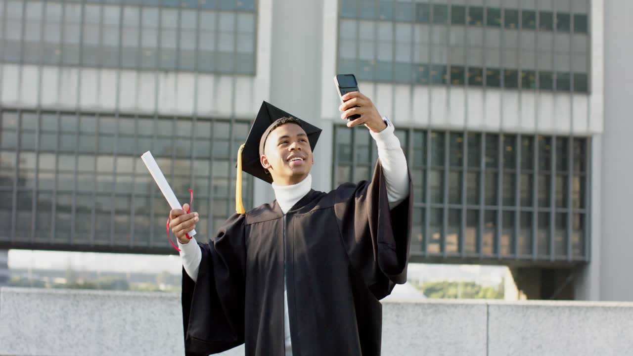 Uomo afroamericano che indossa tocco e toga con diploma, mentre filma la celebrazione con il telefono sul terrazzo