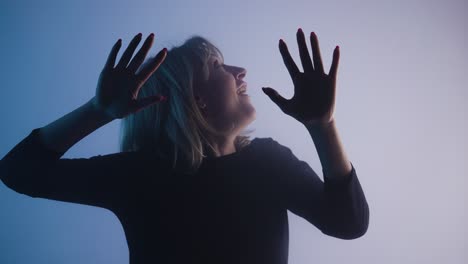 woman looking at glass with wow delight touching it with palms - young woman emerges from smoke, looks at glass with wow delight, touching it with her palms. blonde looks into camera with interest, banging her fists on glass and waving hello. dark studio with soft white blue light. close up