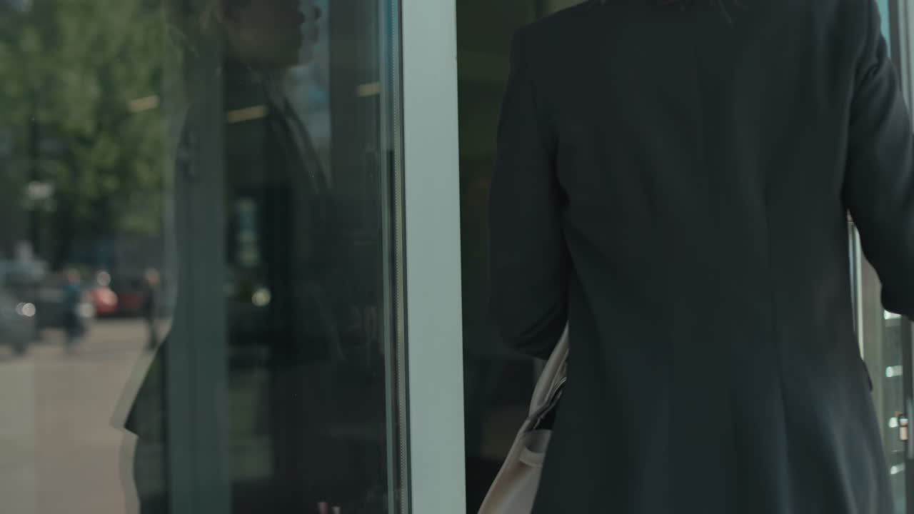 Professional banker opens glass door entering building with her reflection clearly visible on surface, parked cars seen outside in urban setting