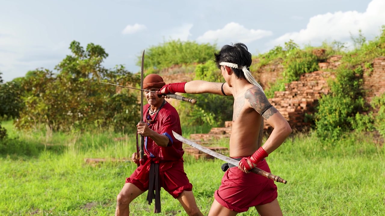 Due guerrieri in abiti tradizionali si esercitano in tecniche di combattimento con la spada all'aperto vicino a rovine storiche in mattoni.