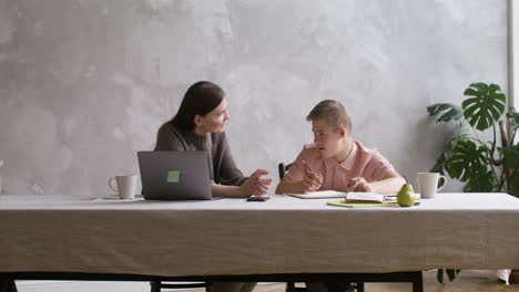 Boy-with-down-syndrome-doing-homeworks-sitting-at-table-in-the-living-room-at-home.-His-mother-helps-him