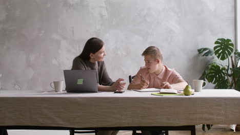 niño con síndrome de down haciendo tareas sentado en la mesa en la sala de estar en casa. su madre lo ayuda