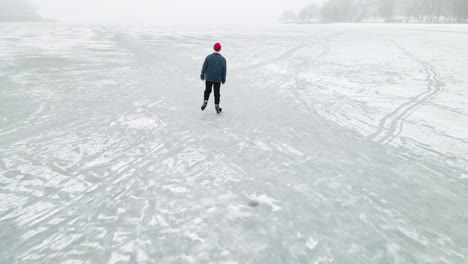 Verfolgungsdrohnenaufnahme-Einer-Person,-Die-Im-Winter-Auf-Einem-Großen-See-Mitten-Im-Wald-In-Einer-Verschneiten-Landschaft-Schlittschuh-Läuft,-Von-Hinten-Gesehen