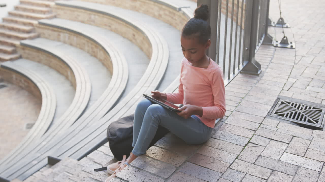 Sitting on steps, African American girl using tablet for studying outside school building