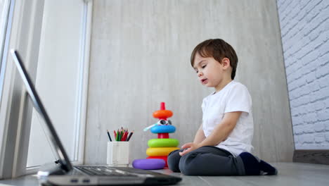 adorable toddler boy in white t-shirt sits near the laptop. cute kid pressing keys and pointing at screen. low angle view.