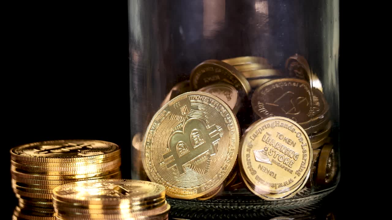 Gold-colored cryptocurrency tokens fall into a glass jar, illuminated by dramatic studio lighting, close-up