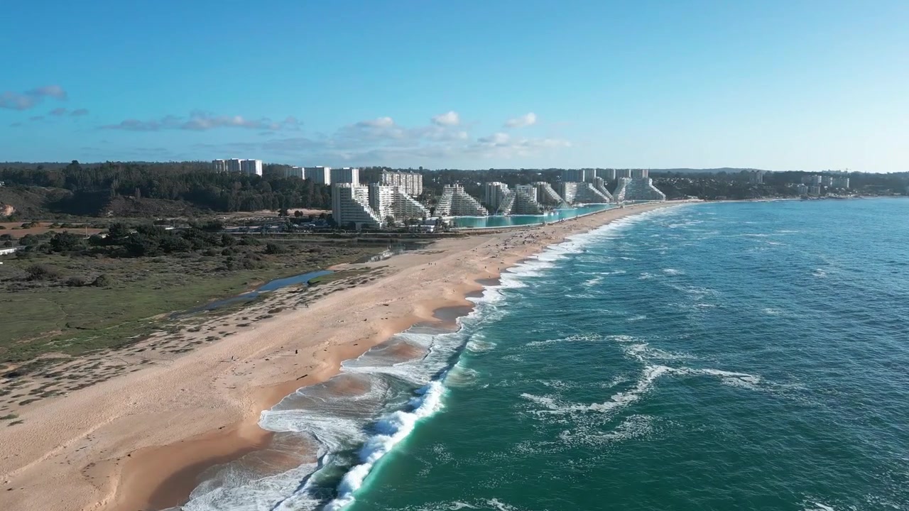 Volo in avanti su una spiaggia sabbiosa incontaminata con onde del Pacifico che si infrangono, rivelando il complesso turistico San Alfonso del Mar e le torri residenziali che si estendono lungo la costa di Algarrobo. Algarrobo, Cile.