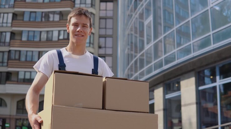 Delivery worker in a white T-shirt and blue overalls holds three boxes on the background of modern high-rise buildings. Express delivery