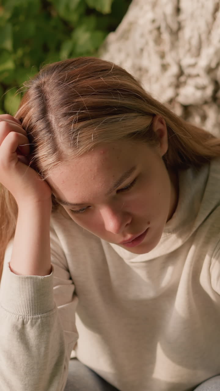 바닥에 앉아있는 젊은 여성은 머리를 왼손에 고, 생각에 빠진 것처럼 보이며, 부드러운 빛이 그녀의 표정을 강조합니다. 배경에는 질감있는 나무 질과 녹색 잎자루가 포함되어 있습니다.