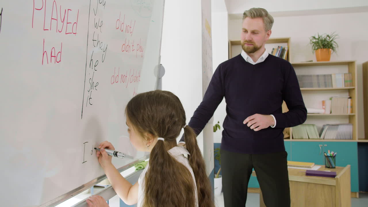 niña pequeña escribiendo una oración pasada simple en la pizarra mientras la maestra la mira