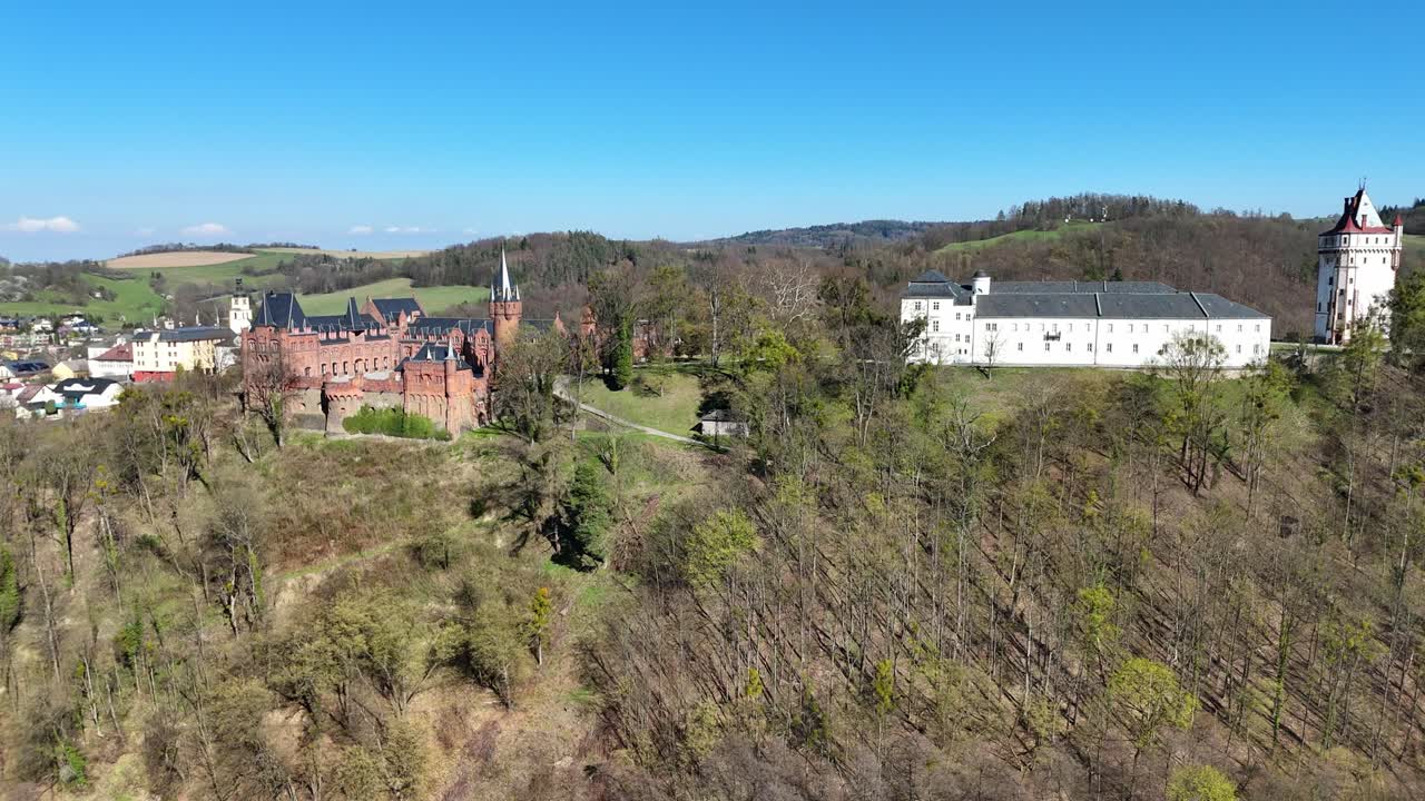 Bevægelse sidelæns til venstre, mens dronen langsomt roterer, afslører slotskomplekset Hradec nad Moravicí med røde og hvide bygninger på en bakke omgivet af træer og naturskønt landskab.