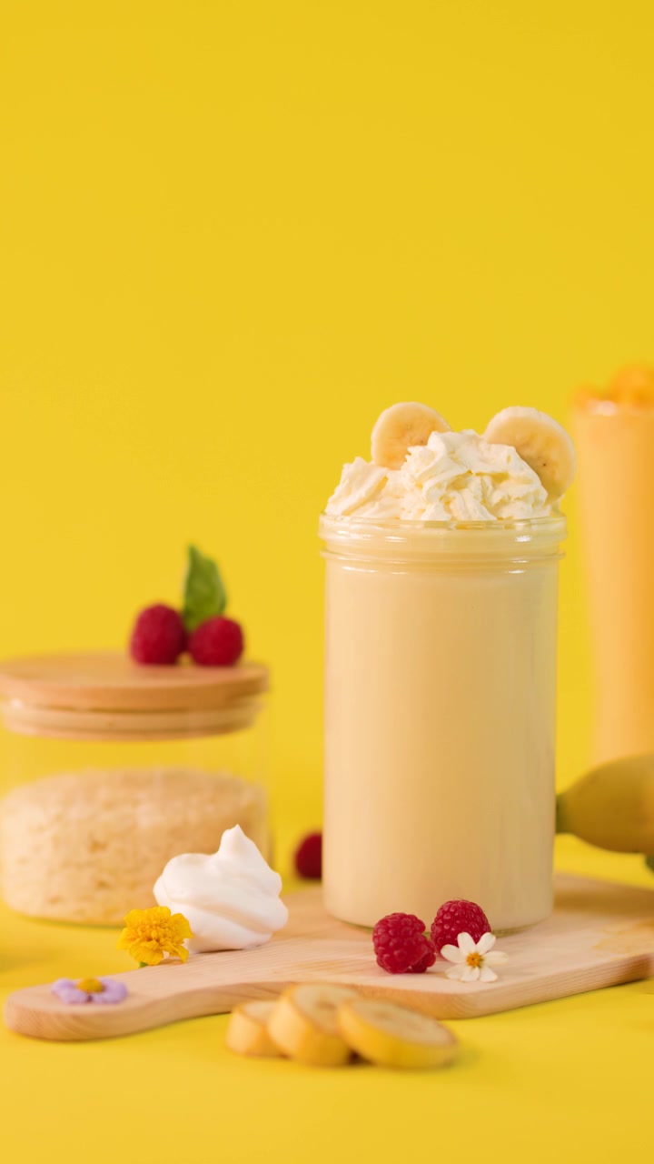 Una mano decora un cremoso frullato di banana con fette di frutta su uno sfondo da studio giallo brillante, telecamera fissa