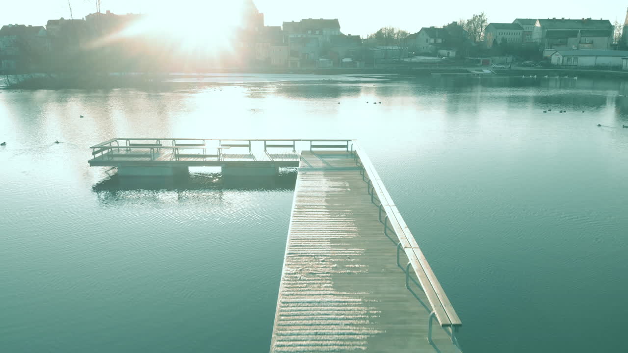 A peaceful flight over an empty pier on the shore of a pond, a water reservoir in a small town, in the background buildings of an old maista,