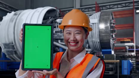 close up of asian female engineer with safety helmet smiling and showing green screen tablet to the camera with airplane engine maintenance conducted