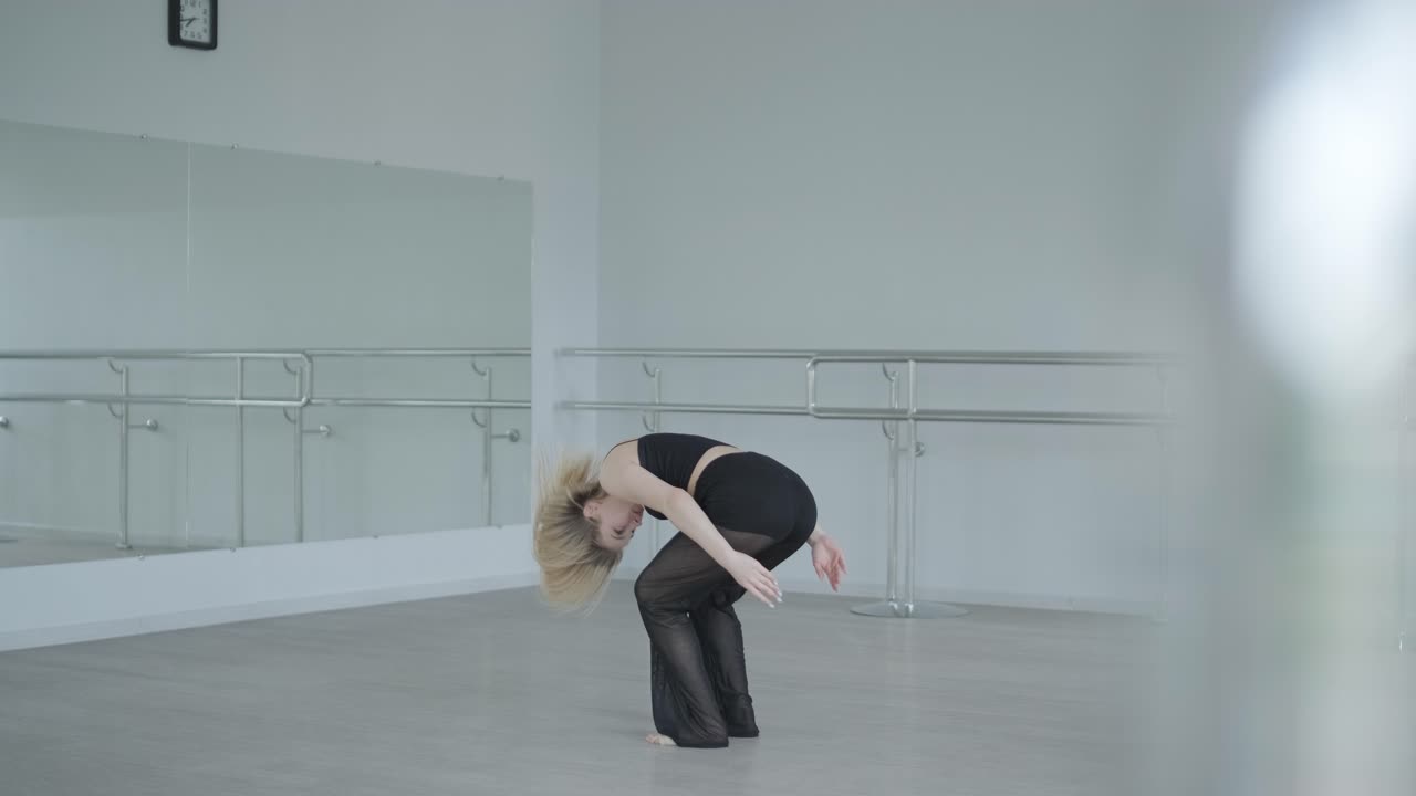 Fit girl in black clothes performing contemporary. Female dancer shows flowing motions, body and hands waves at white room. Modern ballet dance choreography. Young woman is moving smoothly