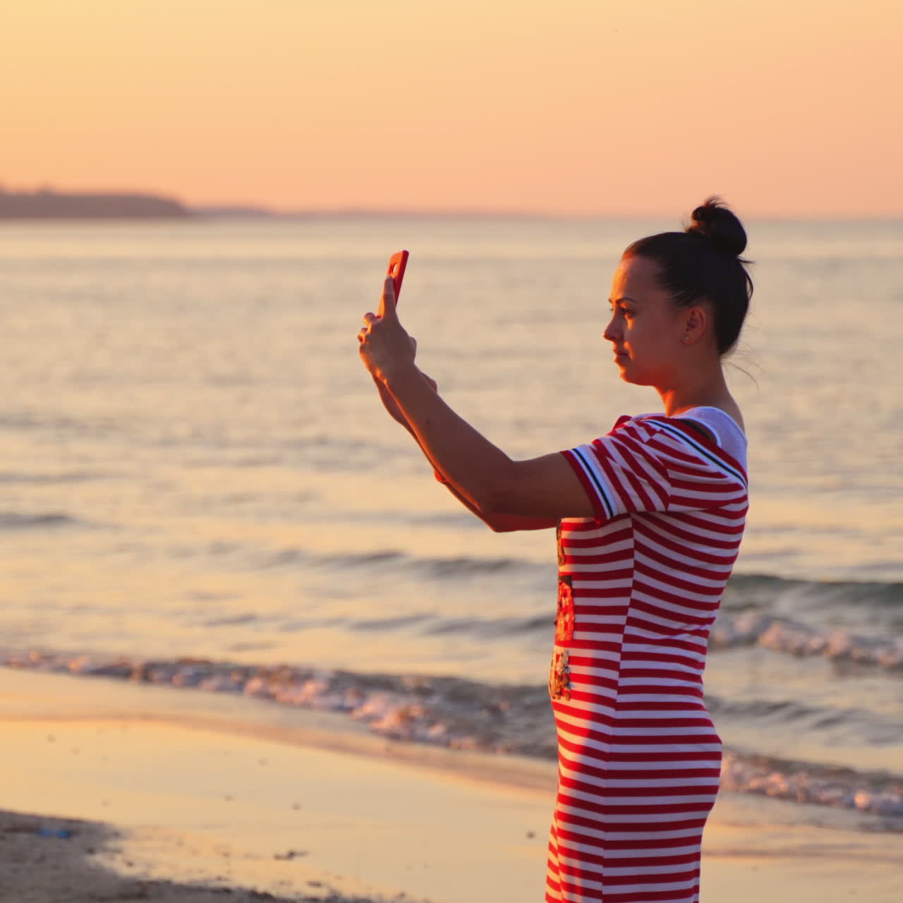 Woman takes picture of beautiful sunset sea with phone. Girl in dress holds smartphone making photo memories of summer vacation with mobile camera.