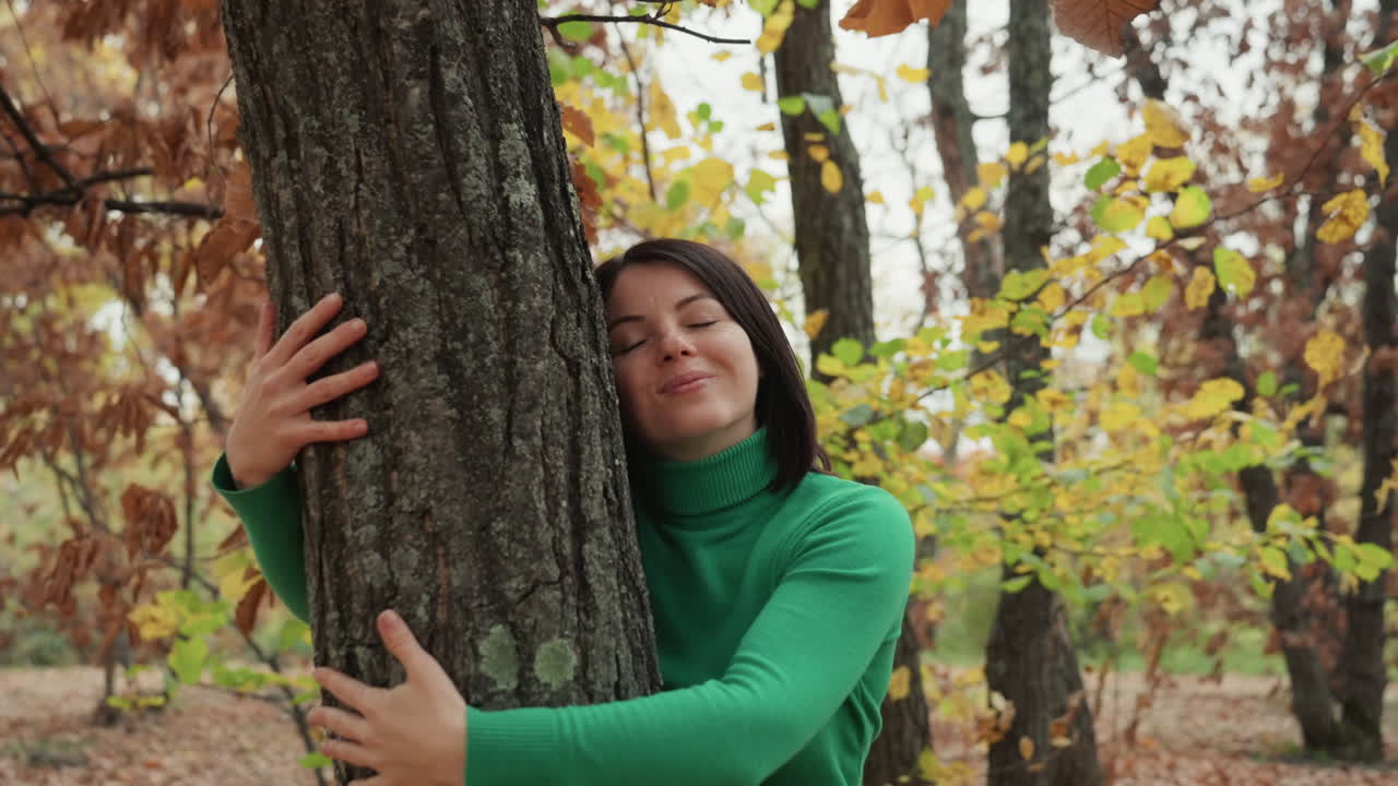 Mujer con un jersey verde explorando un bosque, paseando entre troncos de roble, intentando tocar la corteza, actuando como una botánica que examina el liquen, posando como una fotógrafa de naturaleza, sonriendo con curiosidad divertida