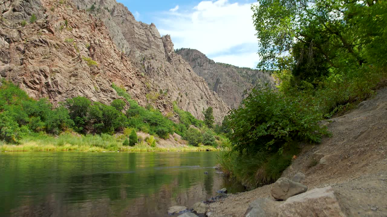 Crystal-clear, emerald-like water flows through Black Canyon of the Gunnison National Park, Colorado. Surrounded by lush green trees, gentle waves ripple from a soft breeze, rocky landscapes.