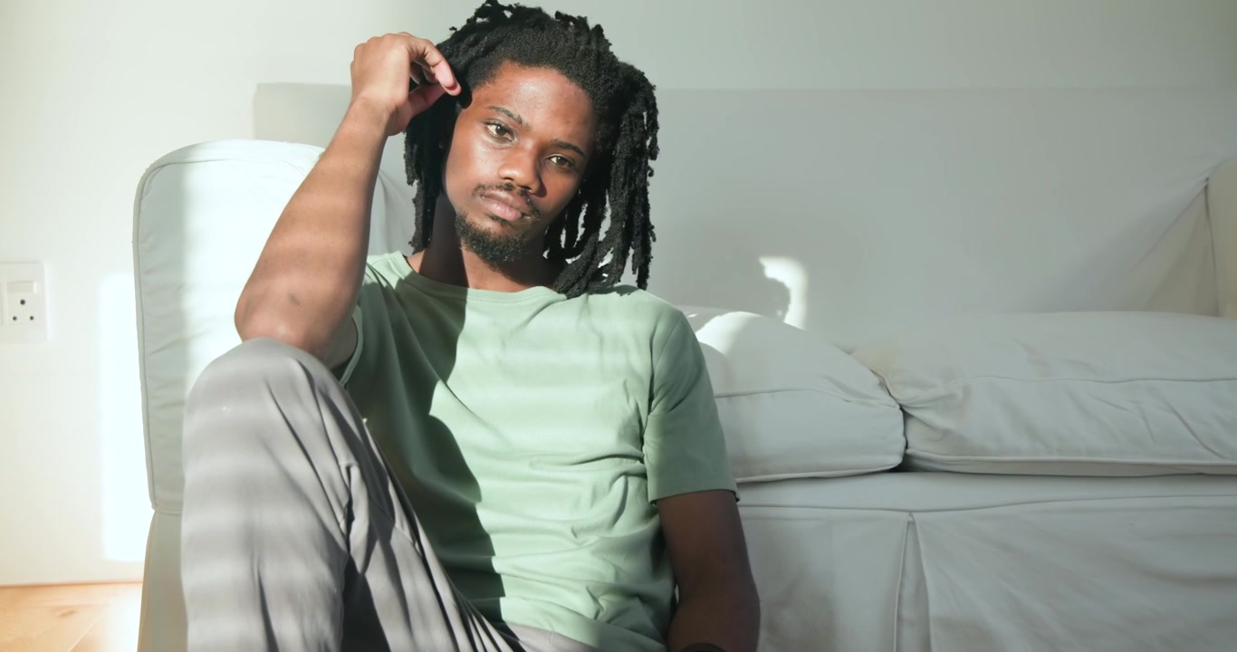 Uomo afroamericano seduto sul pavimento, appoggiato al divano, le tende si muovono, si sistema i capelli, riflettendo