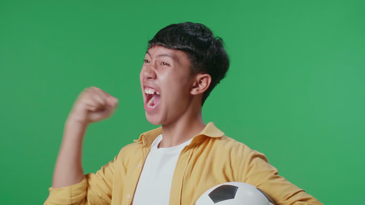 공을 들고 있는 아시아 소년의 가까운 모습. 골을 비명을 지르며 우승팀을 축하하면서 초록색 스크린 배경에서 축구를 응원합니다.