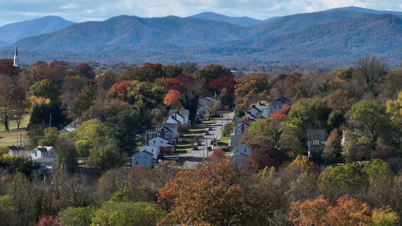 Row of houses between multi-colored trees on hill with mountains in background. Aerial wide shot. American suburb neighborhood with row of houses and church tower. Lynchburg, Virginia