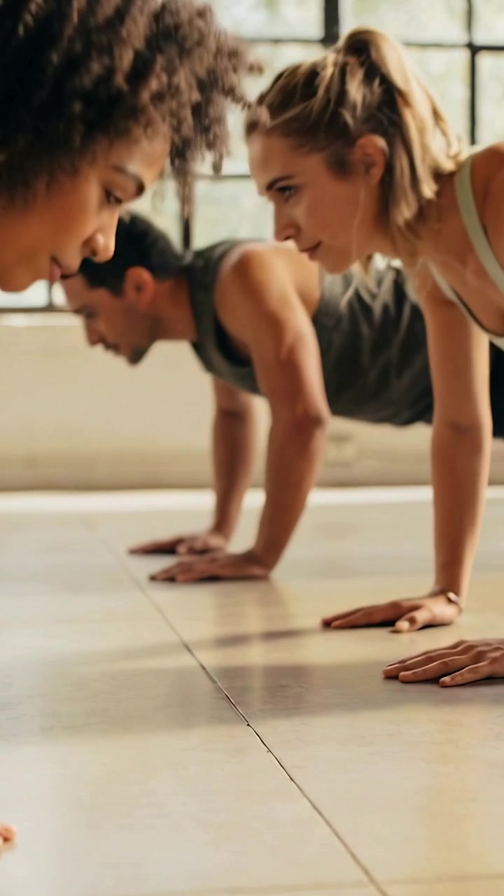 Vertical video: Lowering trio on class cue, doing push-ups in studio by windows in tanks