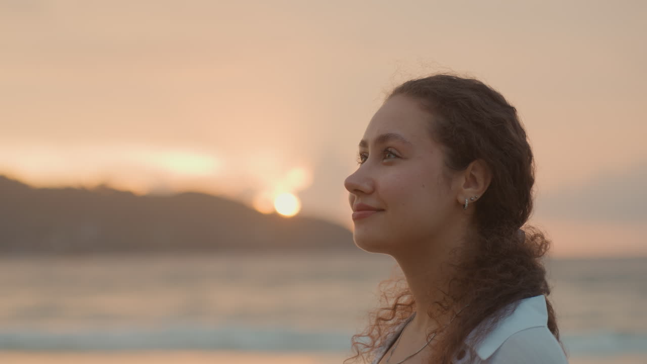 Giovane donna su una spiaggia dell'isola che guarda l'oceano al tramonto, luce soffusa dell'ora d'oro, silhouette di un'isola lontana, profilo laterale contemplativo, capelli ricci fluenti, orecchino discreto, cardigan rilassato, onde leggere.