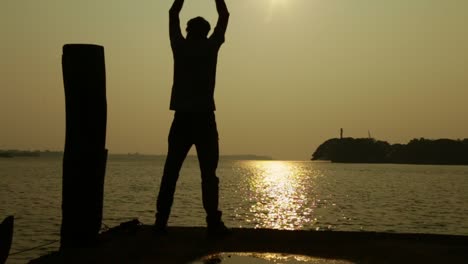 Ein-Mann-Streckt-Bei-Sonnenuntergang-Seine-Arme-Am-Ufer-Aus,-Sonnenuntergang,-Hausboote-Im-Hintergrund,-Silhouette