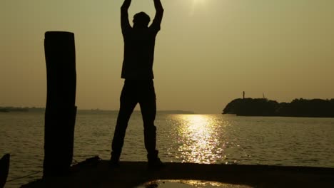 a man stretches his arms by the shore at sunset ,sunset time , houseboats in the background , silhouette