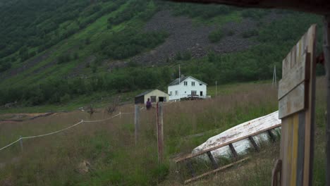 Frau-Geht-Mit-Seinem-Hund-Eine-Grasbewachsene-Straße-Entlang-Zum-Haus-Am-Fuße-Des-Berges-In-Flakstadvag,-Norwegen