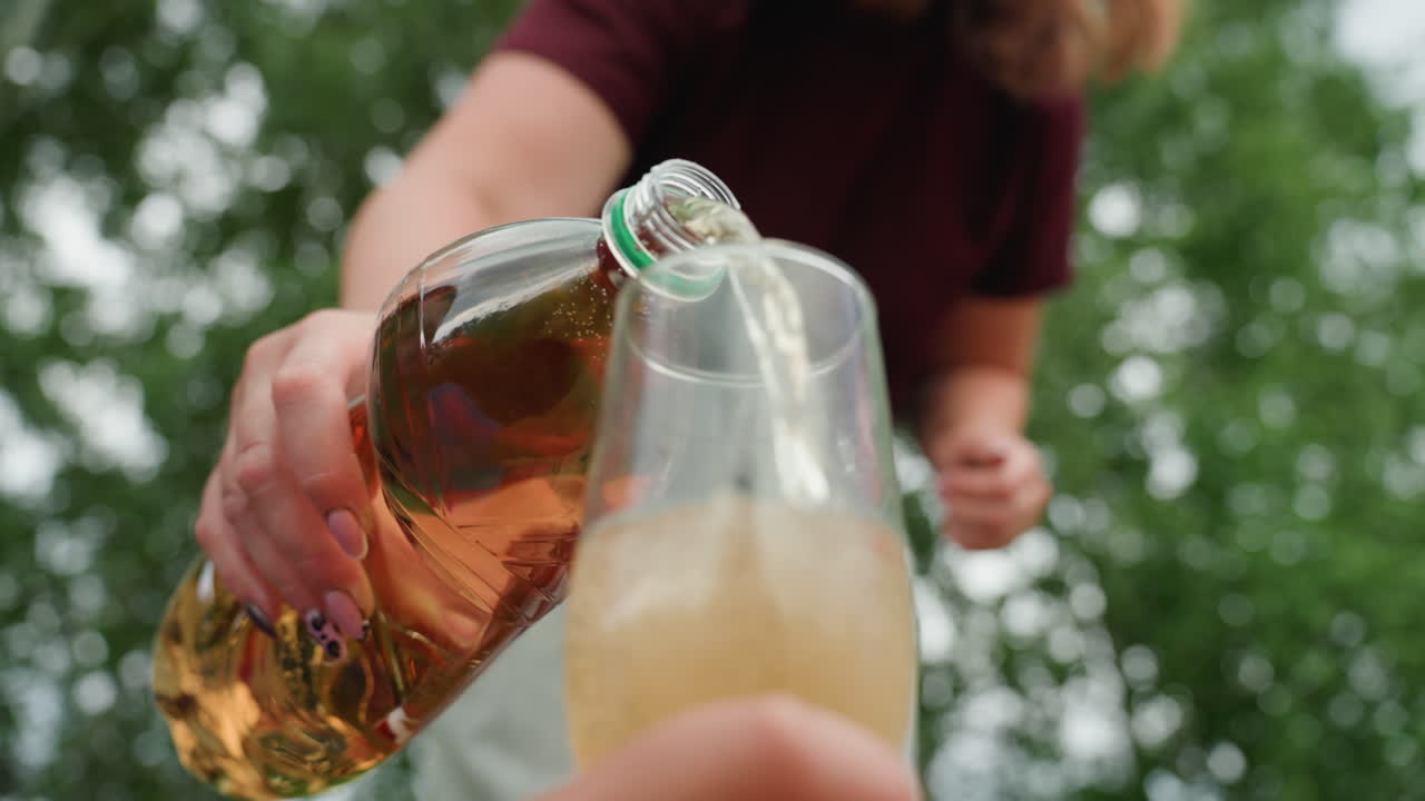 크리미한 거품, 호박색 병, 반짝이는 거품이 있는 맥주를 백인 여성이 잔에 따르고 있어요. 햇빛이 비치는 나무와 뒤뜰 피크닉 분위기, 호스트가 안정된 손으로 음료를 제공하는 클로즈업 야외 사진입니다.