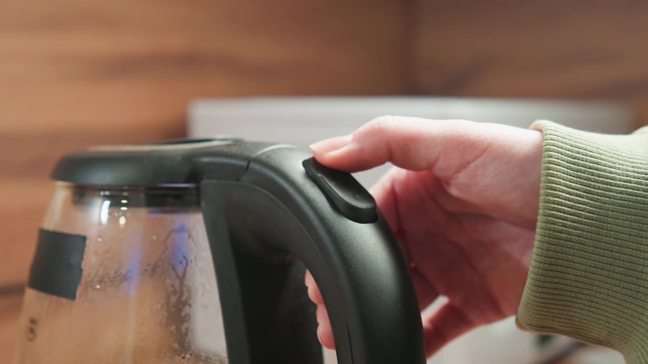 Low-angle close-up of electric kettle handle with finger-operated power switch and tilted glass body, water droplets and condensation visible on surface, showing heating process and pour-ready