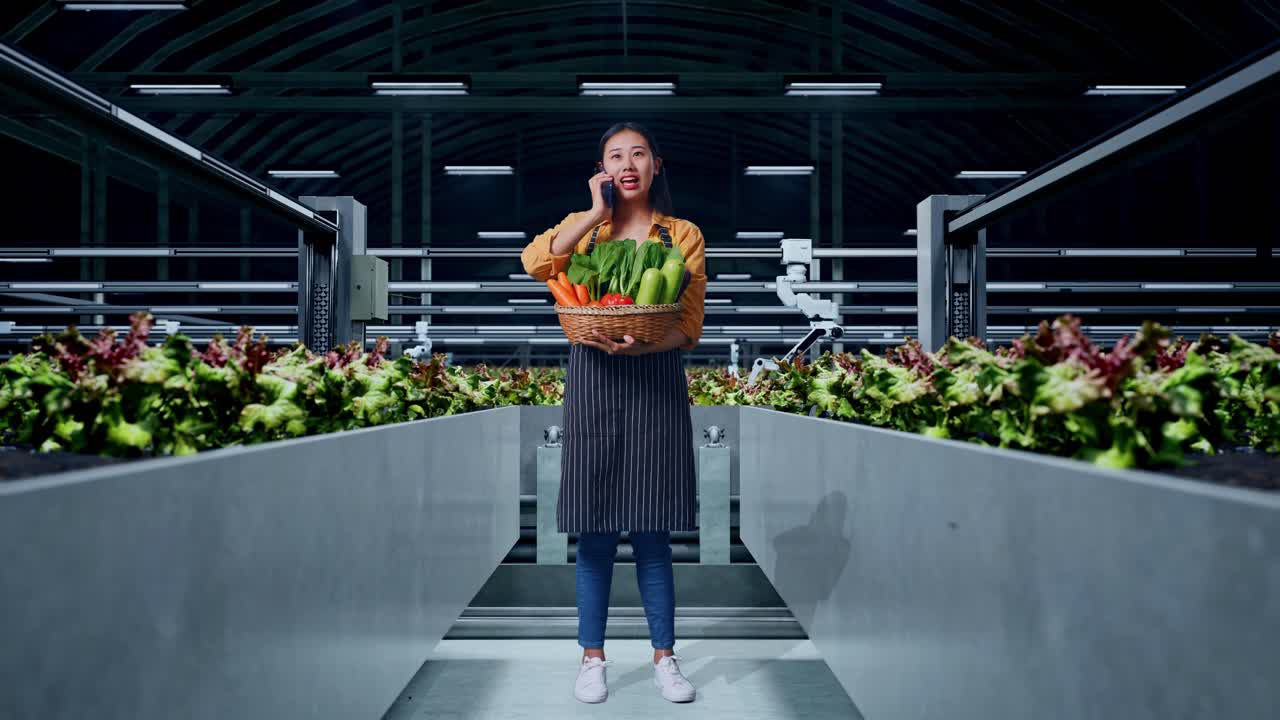 cuerpo lleno de una sonriente agricultora asiática con una canasta de verduras hablando por teléfono móvil en una granja inteligente