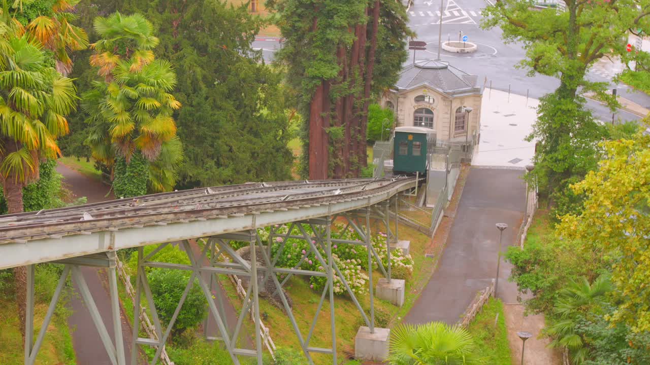 The historic green funicular car climbing the steep railway track in Pau, France, with surrounding lush tropical-style foliage, showcasing a unique method of public transportation