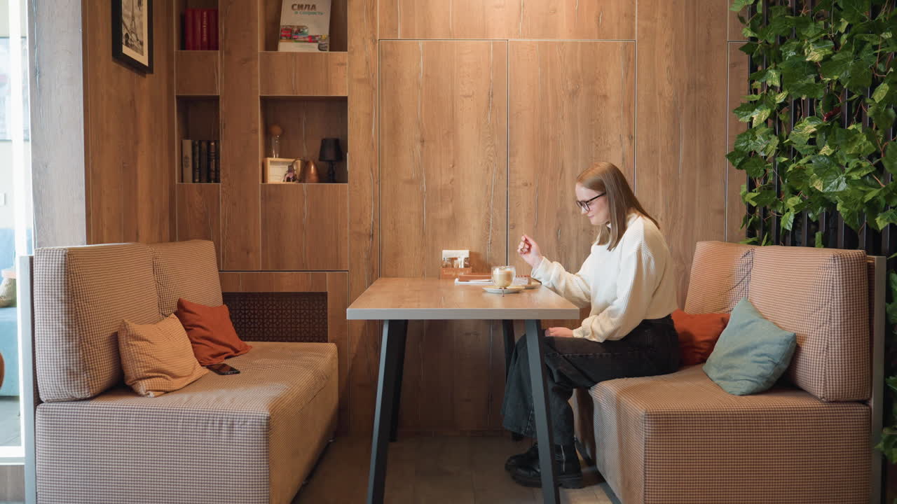 Side view of cheerful woman smiling and clapping hands in excitement before enjoying dessert and coffee in cozy wooden cafe with warm lighting, pillows, bookshelf and greenery around