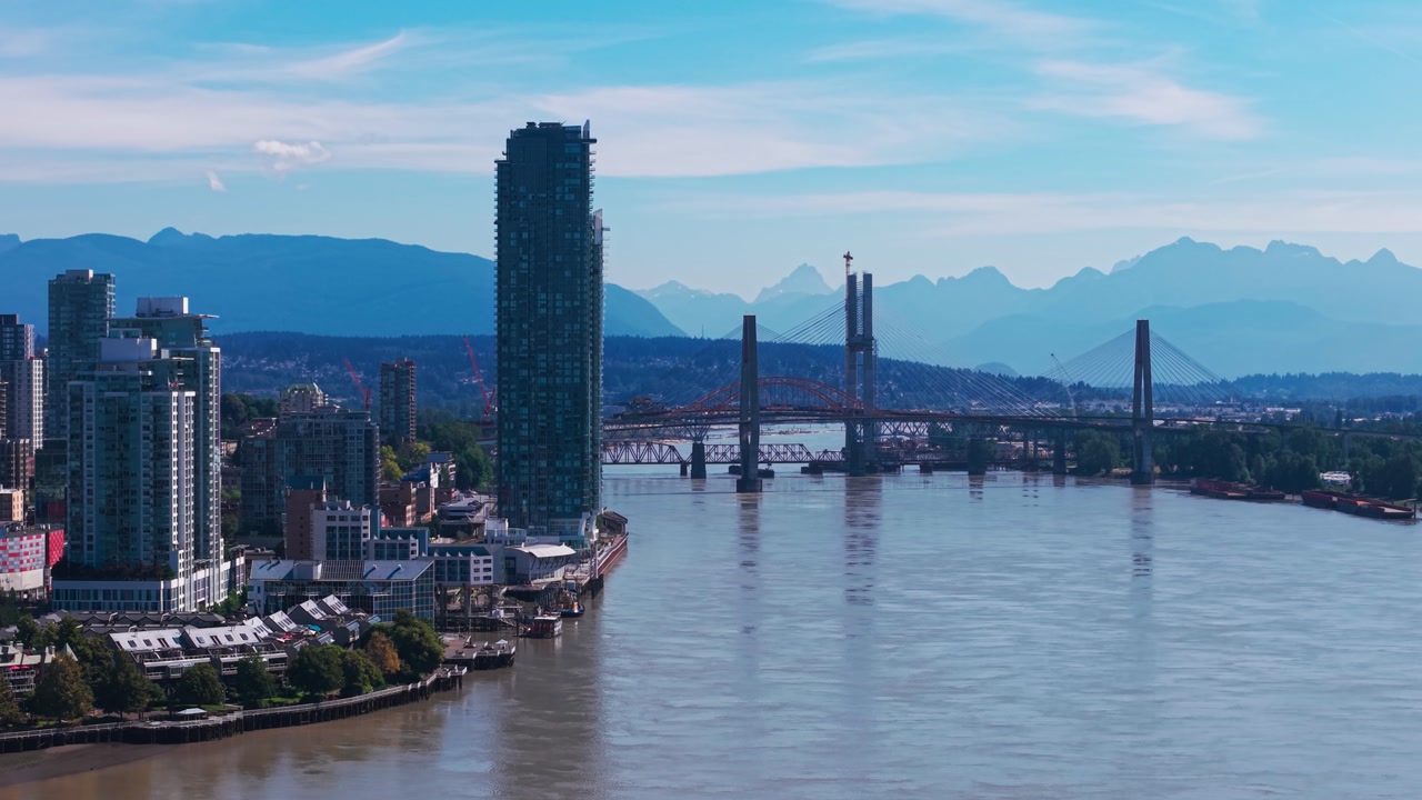 Aerial view of New Westminster skyline featuring SkyBridge crossing the Fraser River, modern high-rises, calm water, and mountains under a clear sk
