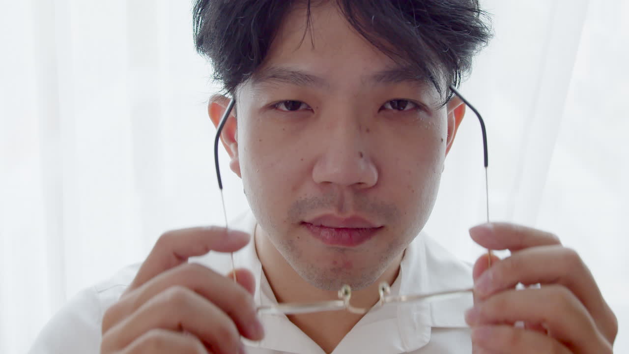 안경을 쓰고 건강한 좋은 머리를 가진 아시아 남자가 카메라를 바라보고 머리를 정리하여 자신을 망쳤습니다.