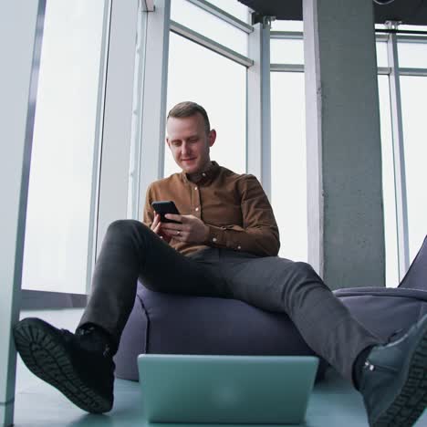 young man uses the mobile phone. businessman is sitting in ottoman with a phone on large windows background. laptop on the floor in office. view from below