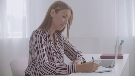 student woman is learning online watching webinar on laptop and making notes in copybook