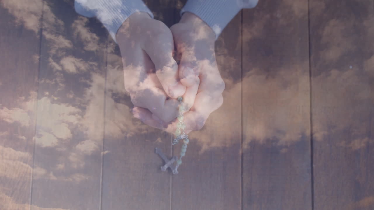 Hands holding rosary with animation of clouds on wooden background