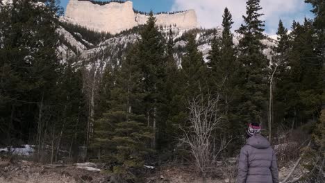 a woman hiker looks at the high cliffs above a snowy forest lake, wide