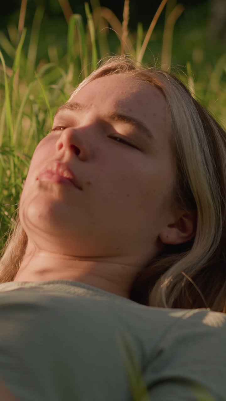 primer plano de una mujer acostada en un campo cubierto de hierba, con su teléfono en la mano, con la luz del sol proyectando un brillo cálido en su cara y la hierba circundante